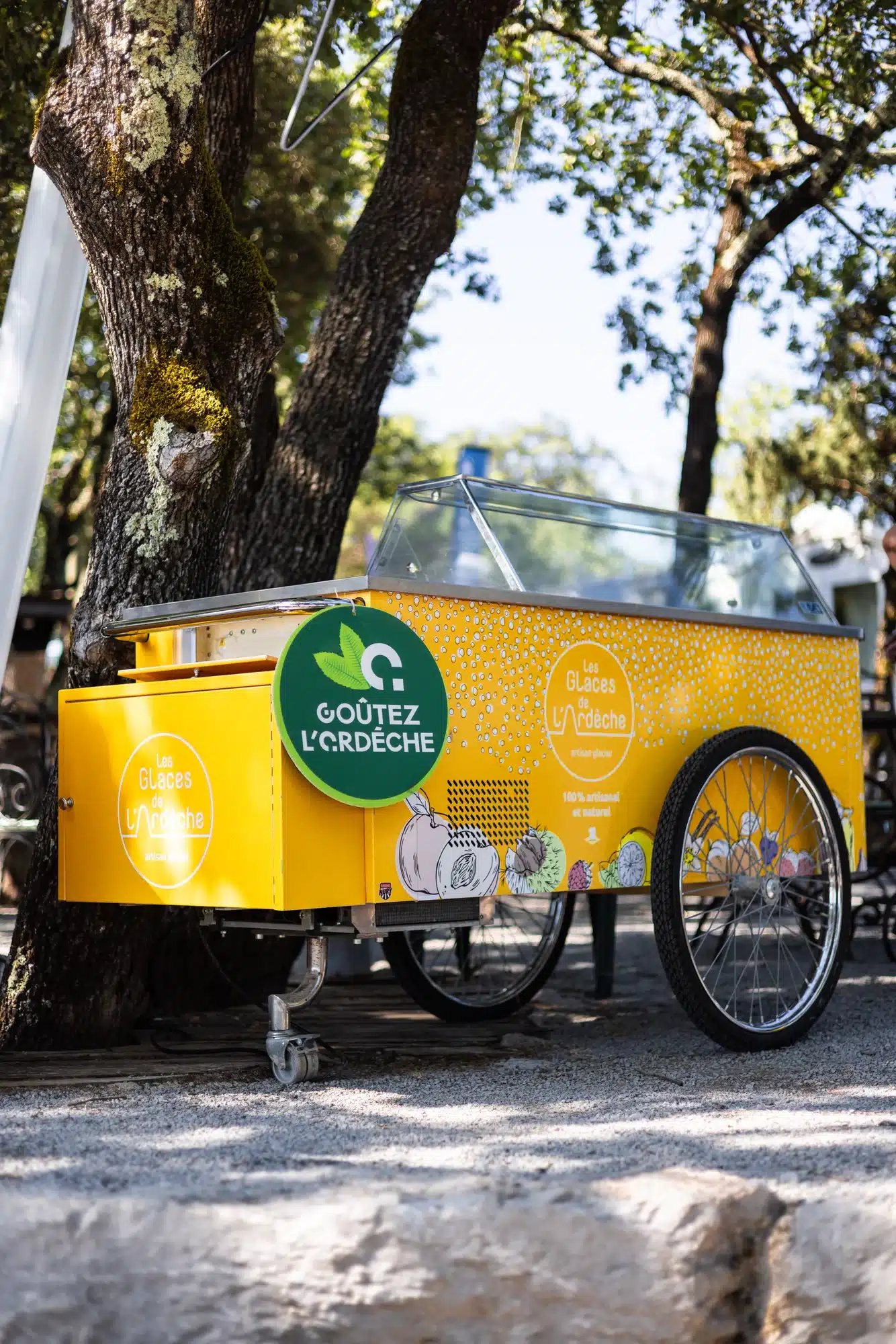 Un chariot jaune vif de glaces artisanales « Les Glaces de l'Ardèche » près d'arbres.