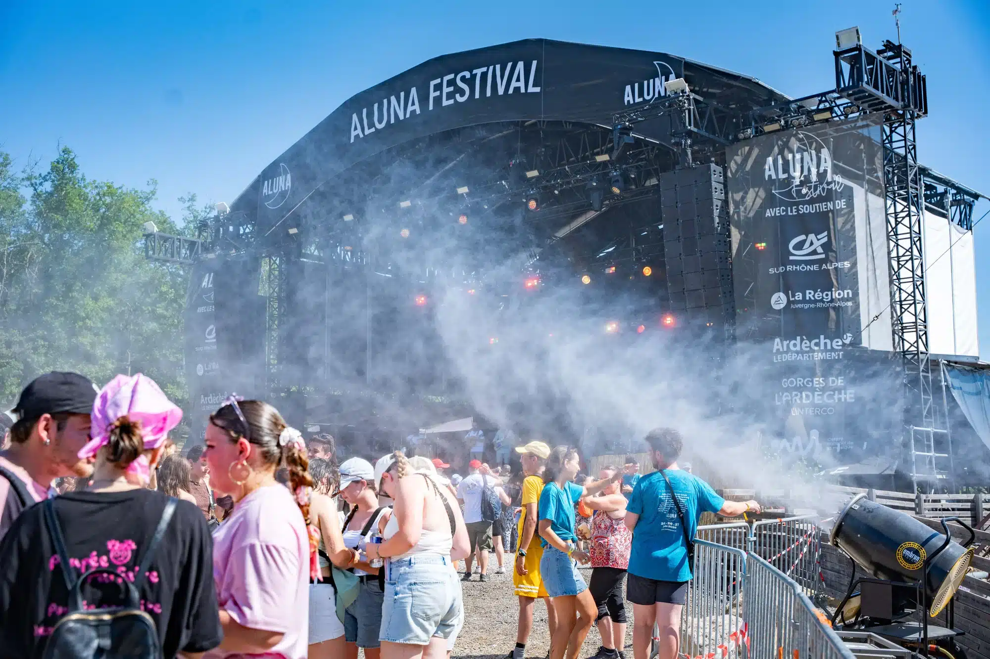 Public rafraîchi par des vaporisateurs devant une scène de festival ensoleillée. Festival Aluna.